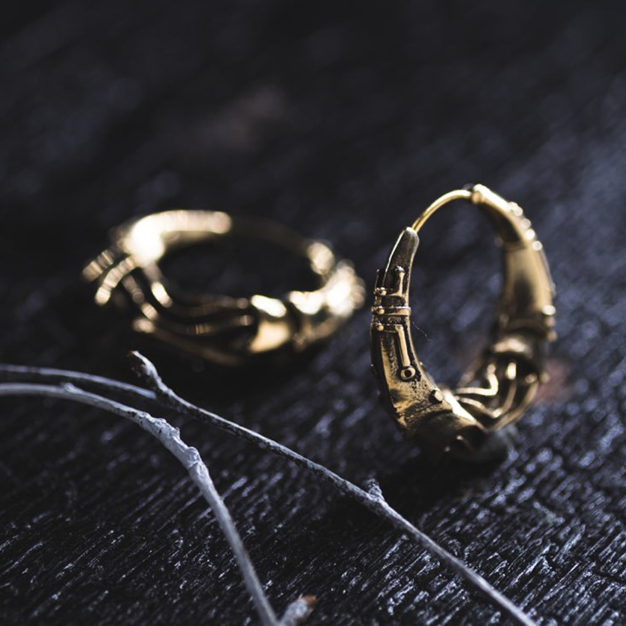A close-up of two intricately designed gold hoop earrings on a dark textured surface. The PROMETHEUS Small Cyborg Hoop Earrings by Kaya Movement feature detailed designs that resemble skeletal fingers. Some thin, dried twigs lie nearby, adding to the moody, post-apocalyptic ambiance of the image.